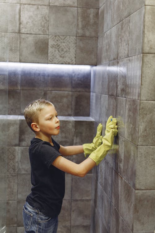 Side view of child with blond hair in yellow rubber gloves brushing tile wall in bathroom