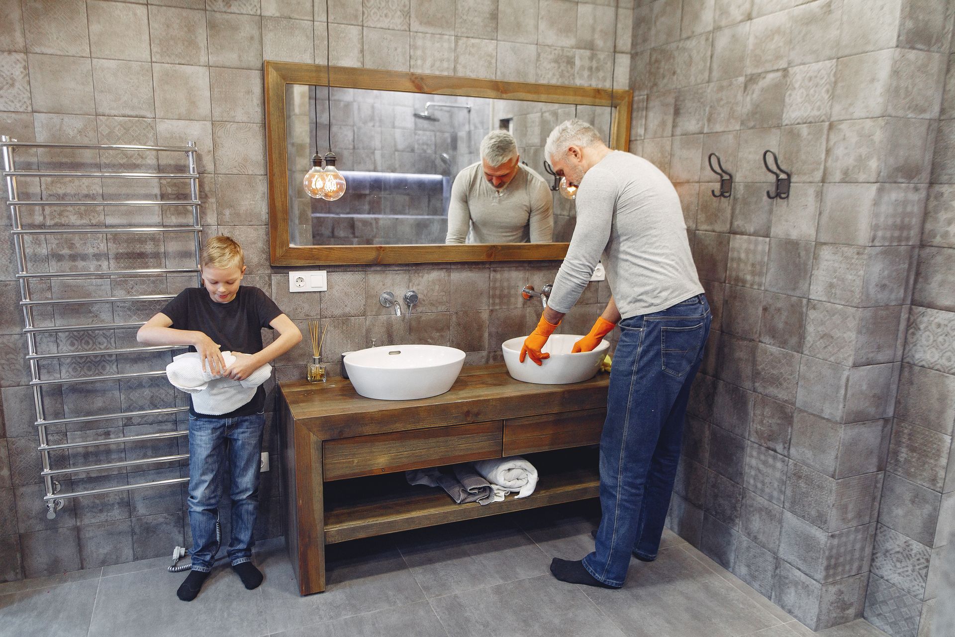 Grey haired dad with beard in orange rubber gloves and small boy cleaning bathroom together