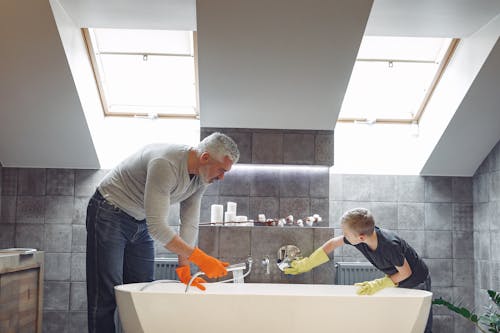 Free Side view of grey haired dad with beard in orange rubber gloves cleaning bathroom together with son Stock Photo
