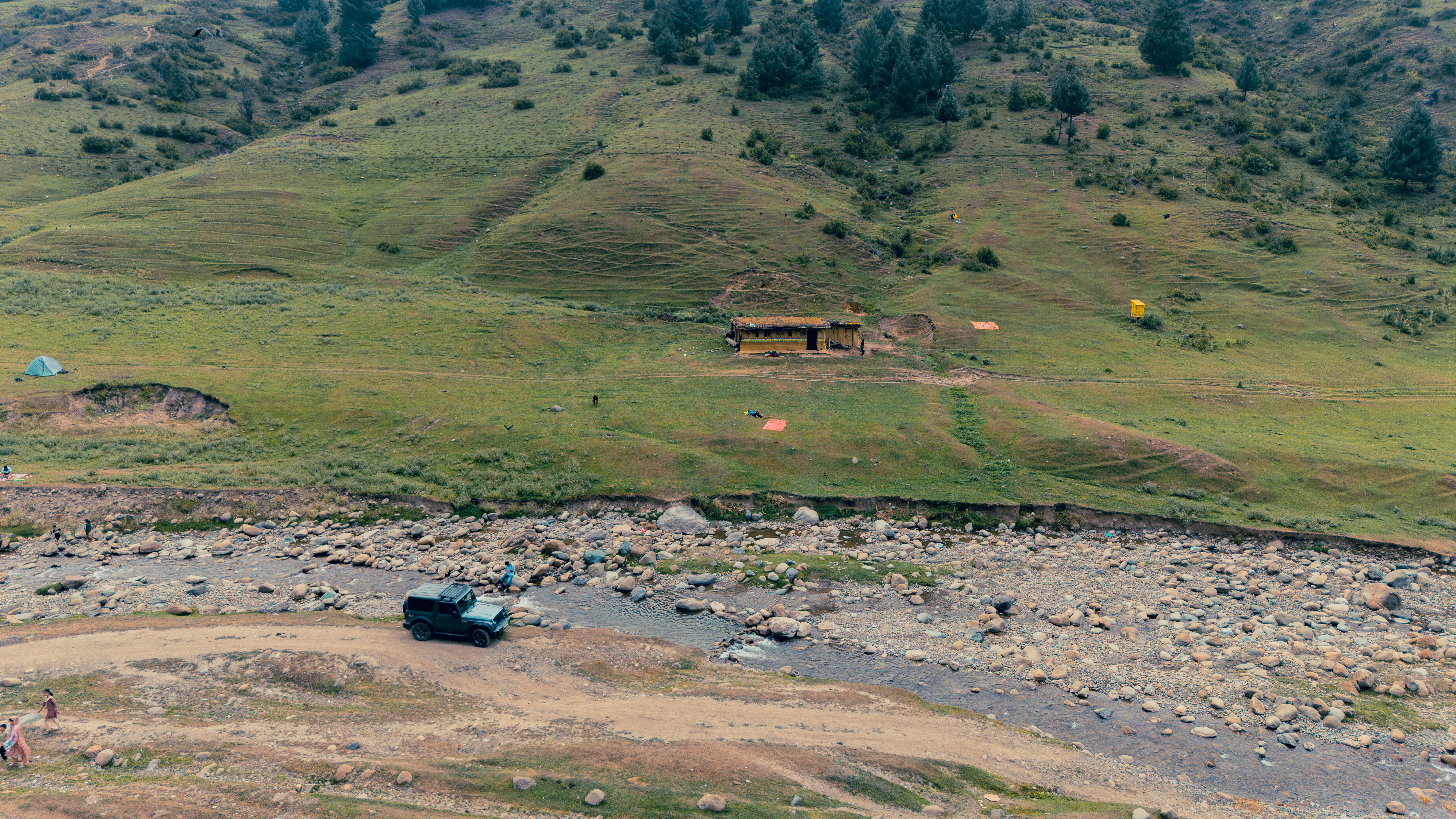 Aerial view of a serene hillside landscape with a stream, vehicle, and tents in Kashmir.