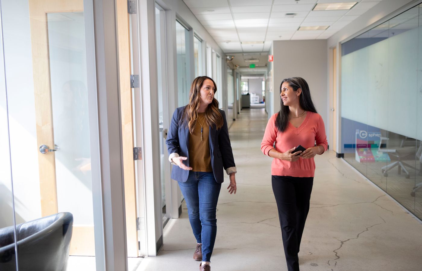 Two professional women engaged in conversation while walking through a modern office corridor.