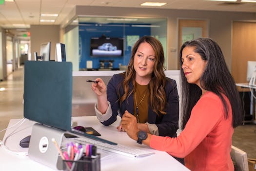 Two professional women discussing project details together in a modern office environment.