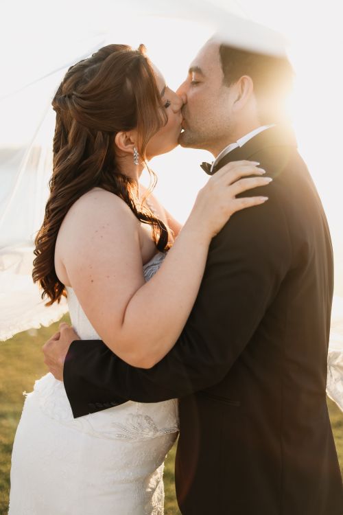 Free A bride and groom share a romantic kiss outdoors during sunset, creating a dreamy moment. Stock Photo