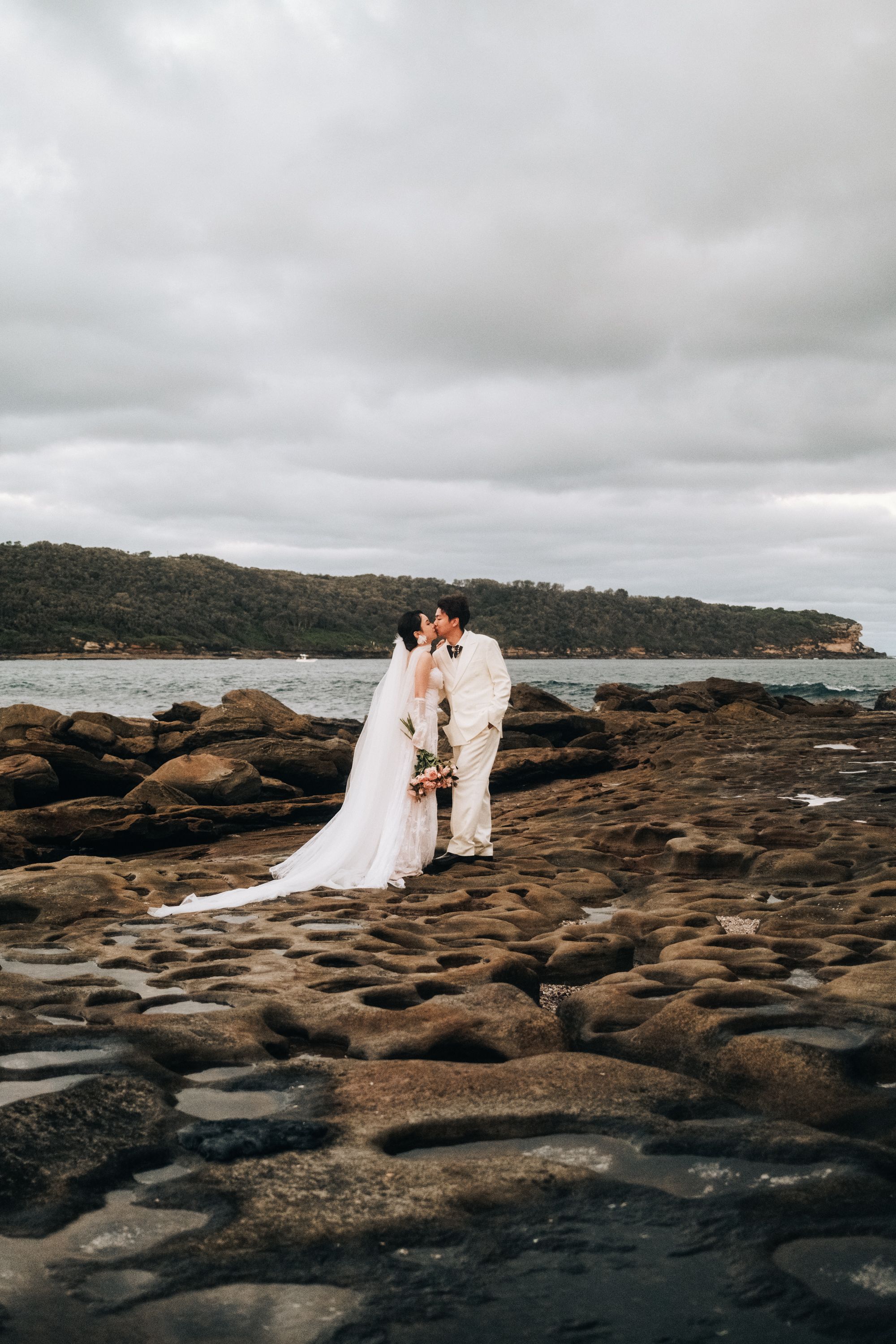 Bride and groom embrace on picturesque rocky shoreline in Sydney, Australia, under a cloudy sky.