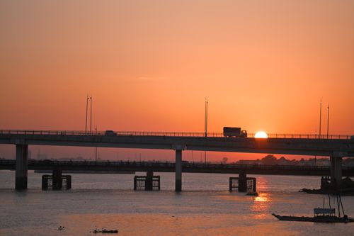 Free A truck silhouetted against a vibrant sunset crossing a bridge over calm waters. Stock Photo