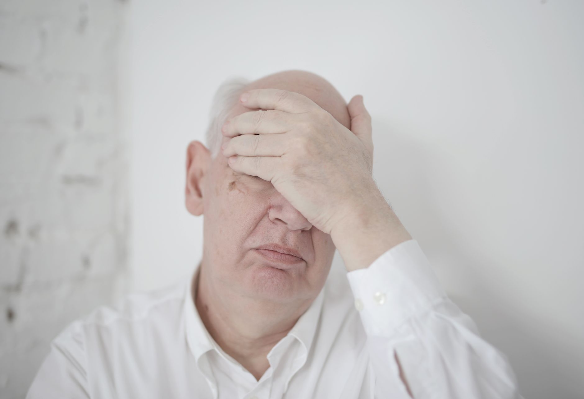 Disappointed senior bald man in white shirt covering face with hand while sitting near wall in light room