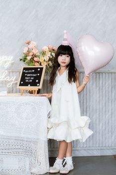 Young girl in white dress holding heart-shaped balloon at birthday party indoors.