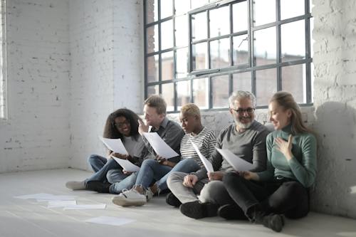 Free Smiling diverse coworkers in casual clothes sitting with crossed legs with sheet of paper leaning on brick wall near fenced window in daytime Stock Photo