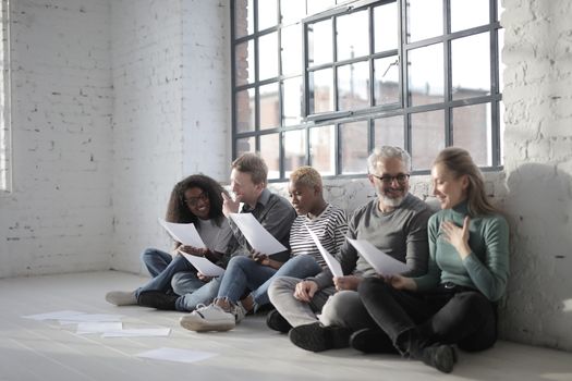 Smiling diverse coworkers in casual clothes sitting with crossed legs with sheet of paper leaning on brick wall near fenced window in daytime