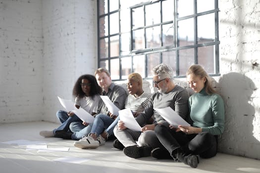 Group of contemporary diverse coworkers in casual wear with papers sitting with crossed legs on ceramic floor near fenced window in afternoon
