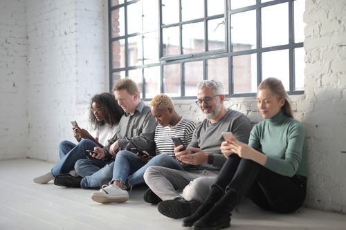 Free Smiling coworkers of different ages in casual outfit browsing internet on cellphones while sitting with crossed legs near brick wall and fenced window during daytime Stock Photo