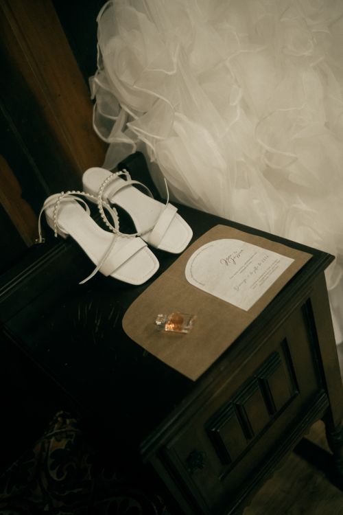 Free A close-up of wedding details including white bridal shoes, dress, and invitation on a vintage nightstand. Stock Photo