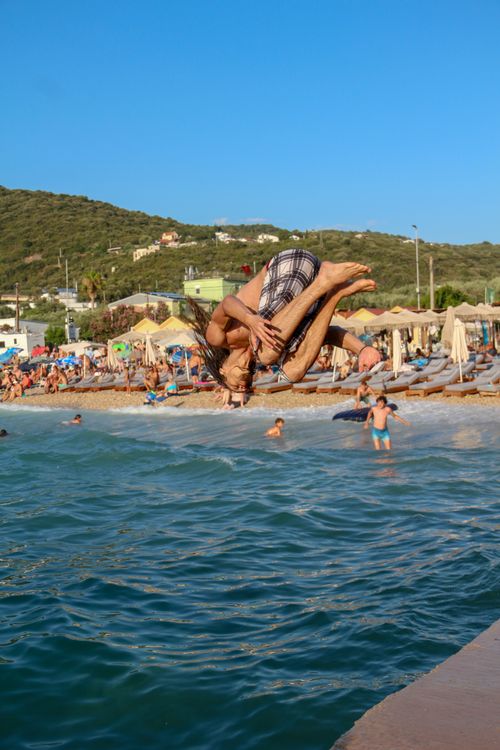 Free A man doing an aerial flip near the water at a sunny beach with people relaxing in the background. Stock Photo