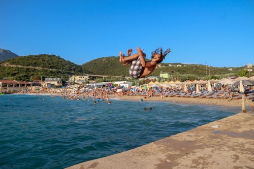 Free A man performs an aerial backflip into the water on a sunny beach filled with people relaxing and enjoying the day. Stock Photo