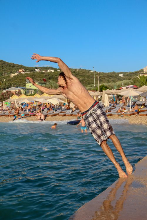 Free A young man performs a diving leap into the clear blue waters at a sunny beach filled with people enjoying the summer. Stock Photo