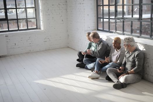 From above of diverse colleagues in casual wear browsing internet on cellphones while sitting with crossed legs near big fenced window in afternoon