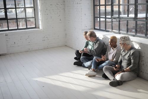 Free From above of diverse colleagues in casual wear browsing internet on cellphones while sitting with crossed legs near big fenced window in afternoon Stock Photo