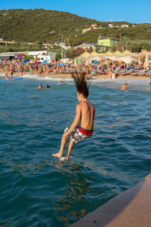 Free A young man with wet hair jumps into the sea from a pier on a sunny day at a busy beach. Stock Photo