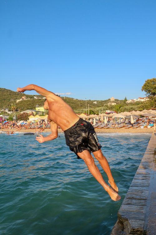 Free A young man in swim shorts diving into the clear blue sea by a crowded beach. Stock Photo