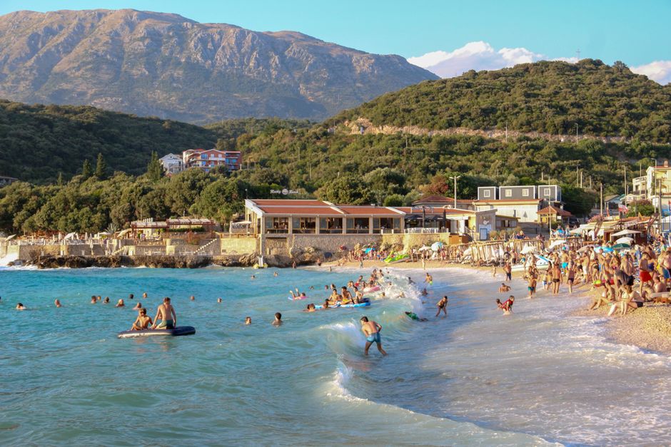 A lively beach scene with people swimming and relaxing against a mountainous backdrop on a sunny day.