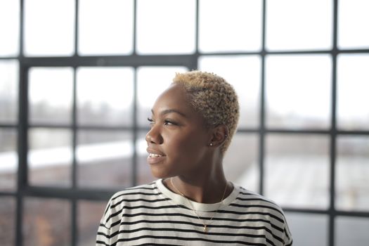 A young woman with short hair gazes thoughtfully out of a window, framed by soft natural light indoors.