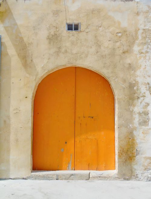 Free A vibrant orange door set in a rustic weathered archway under sunlight. Stock Photo
