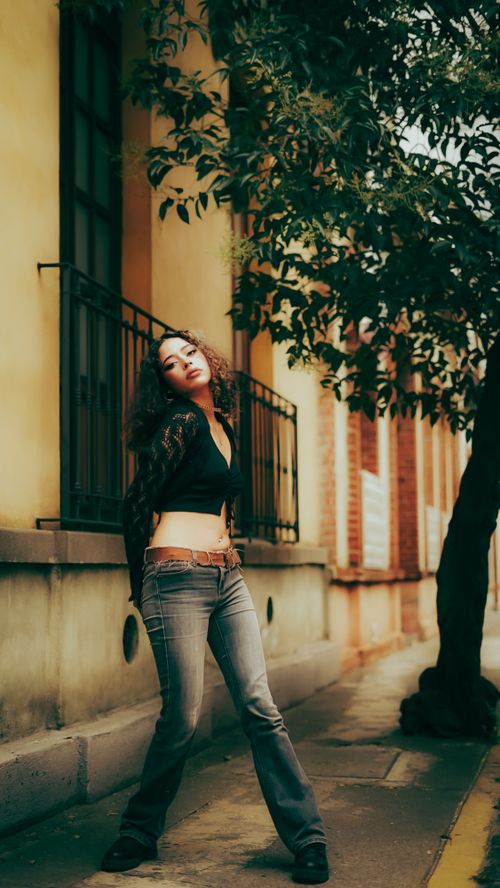 Free Young woman posing confidently in a city street, surrounded by warm urban architecture and trees. Stock Photo