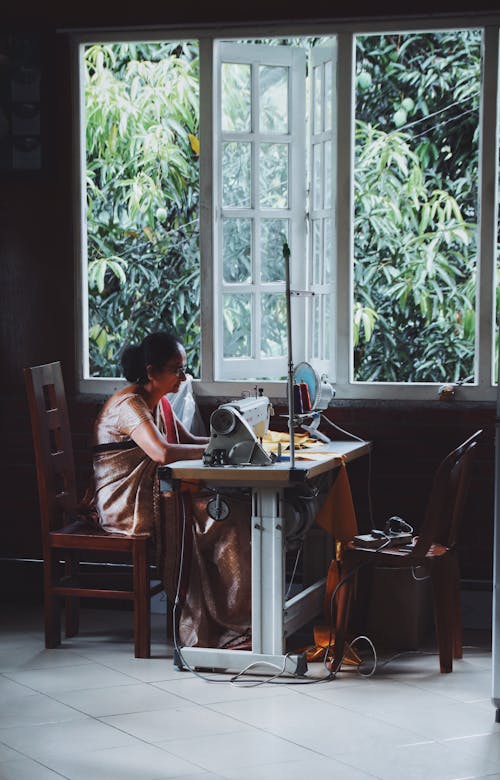 Side view full length focused Indian female tailor wearing traditional sari sitting at table with sewing machine against opened window and working on new garment