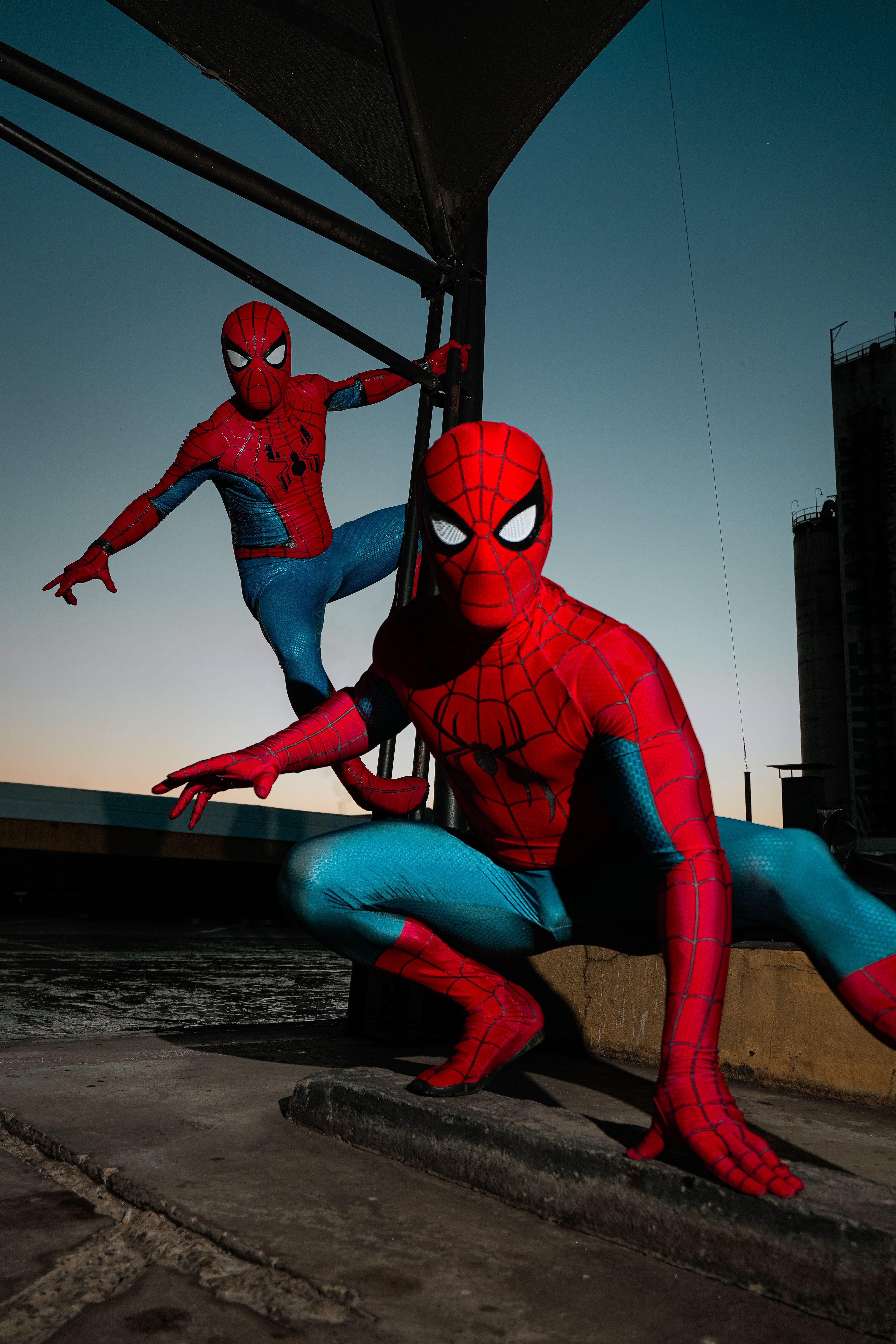 Two individuals in Spider-Man costumes strike dynamic poses outdoors at twilight.