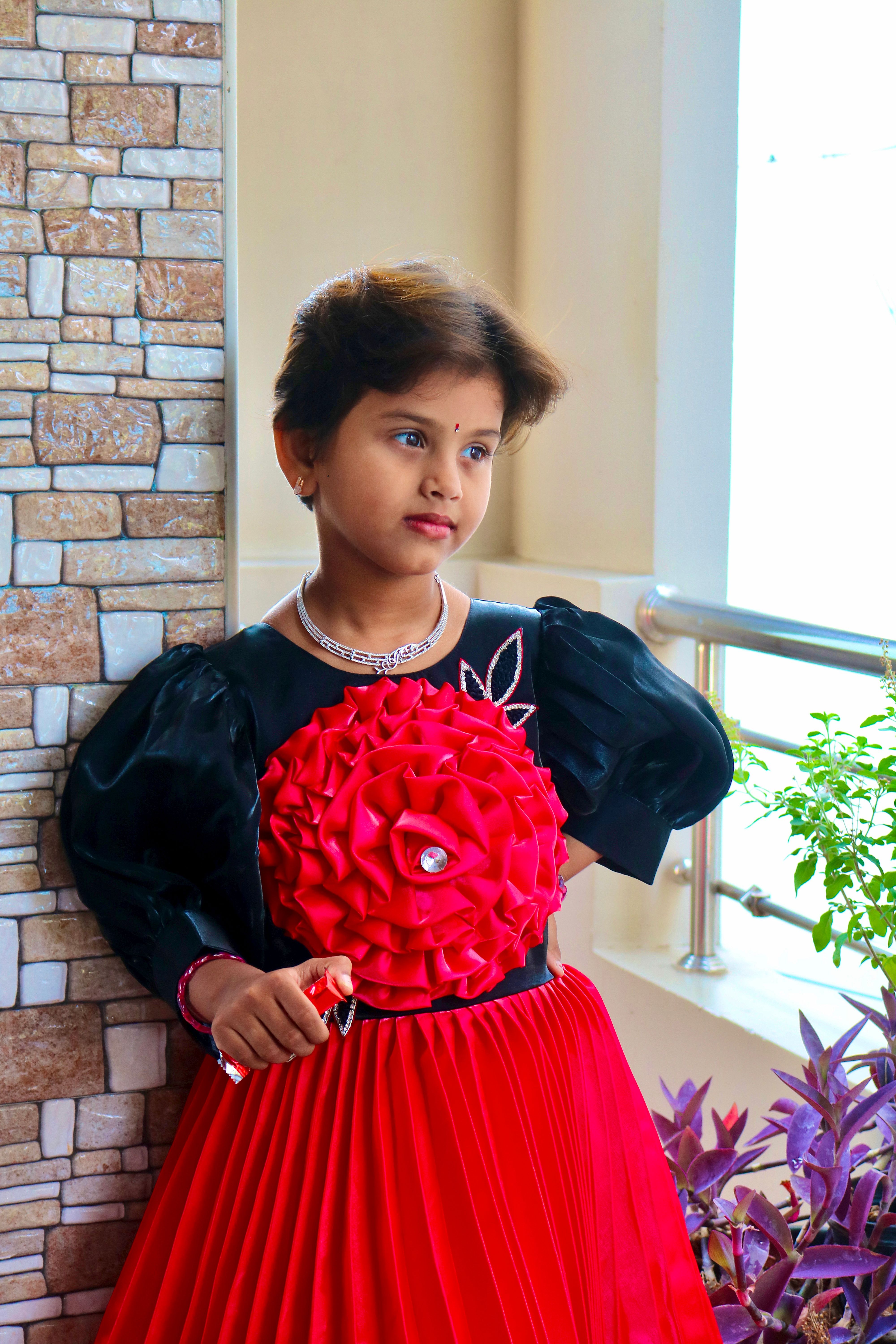 A young girl in a red and black dress poses indoors by a window, exuding elegance.