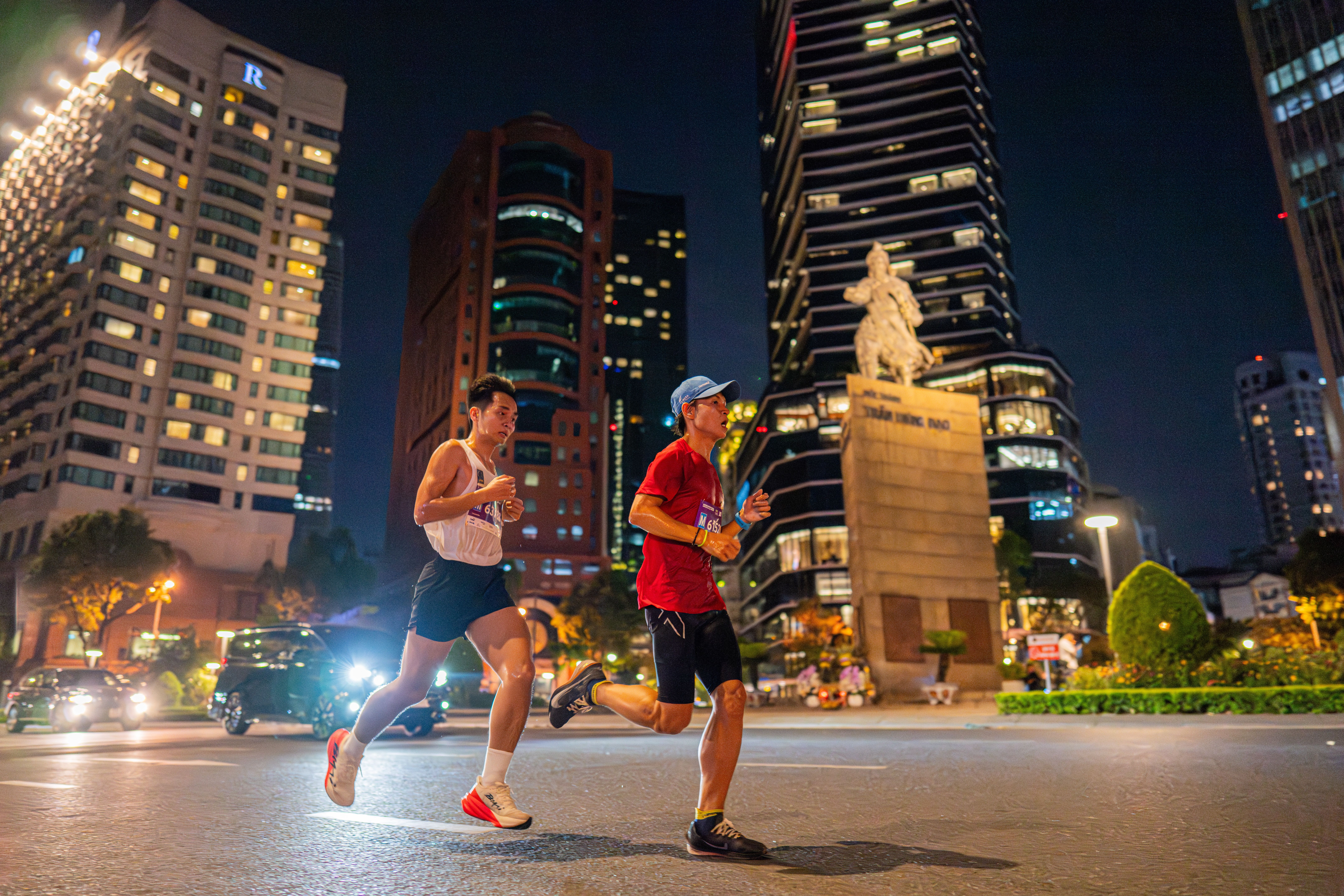 Two men running at night in a bustling city with illuminated buildings and a statue.