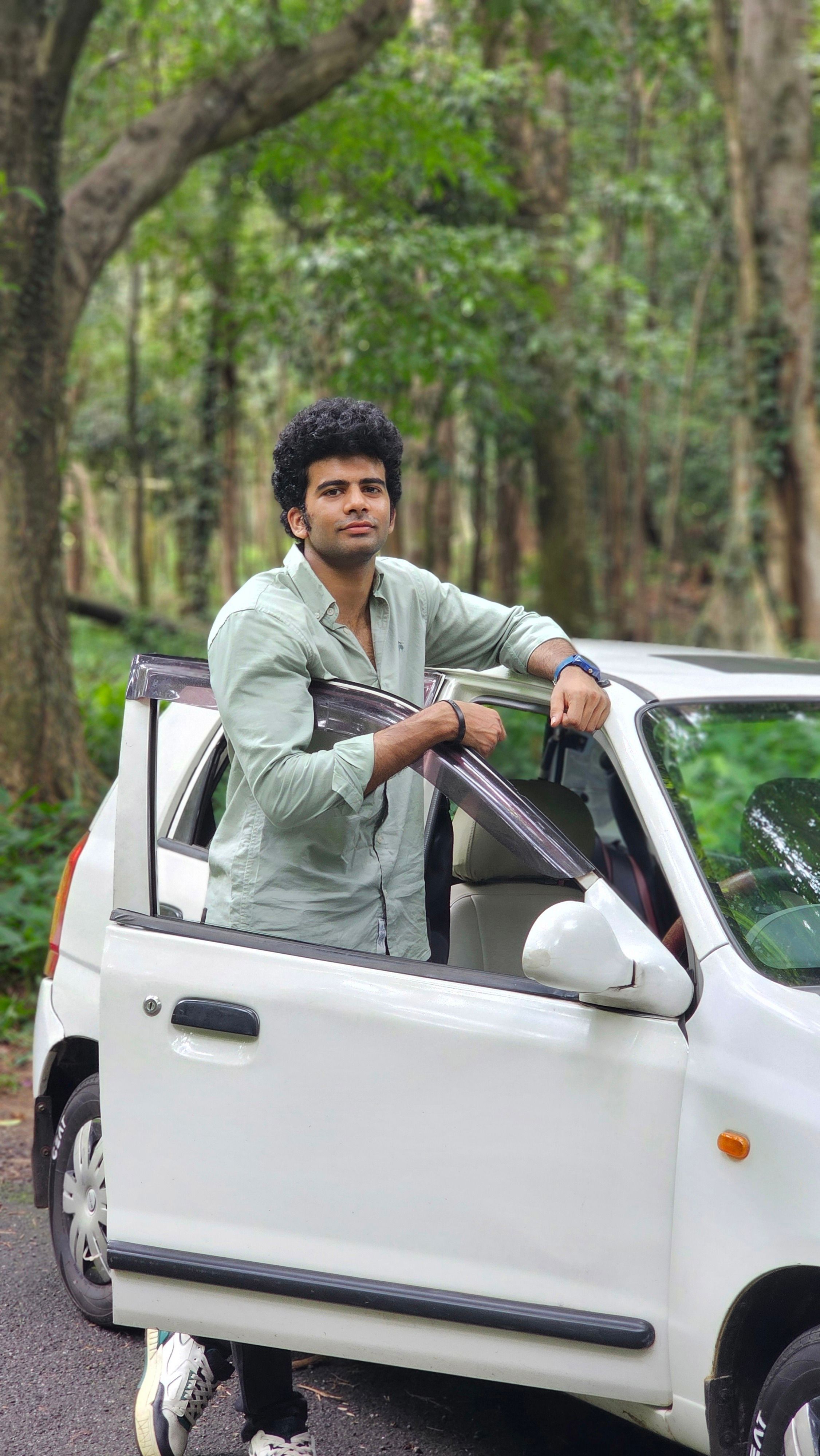 A young man stands confidently next to a white car on a forest road, creating an adventure vibe.