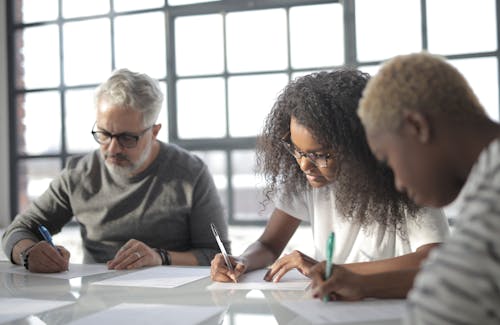 Free Serious multiethnic colleagues in casual wear writing on papers with pen during teamwork while sitting at table in light office behind fenced window Stock Photo