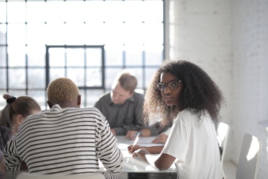 Serious African American female worker writing with pen on sheet of paper while sitting at table near diverse partners and looking at camera in sunlight