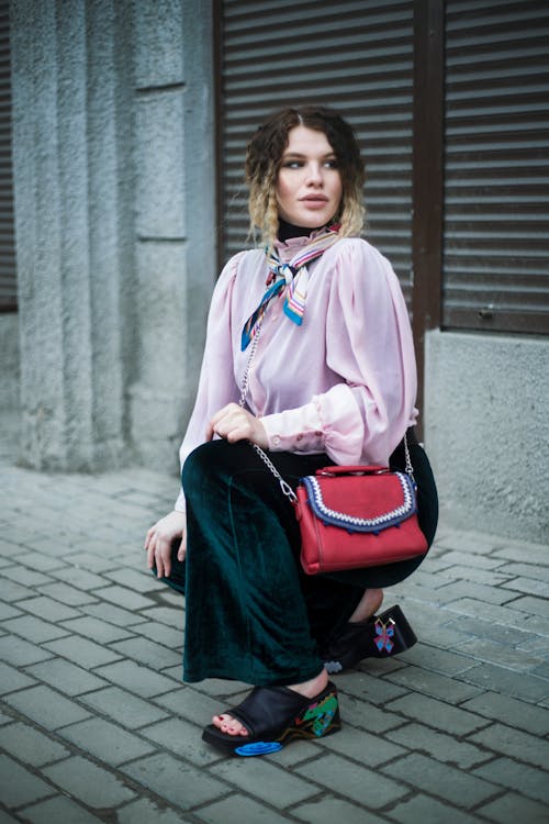 Full body trendy gorgeous female in pink blouse and classy black pants hunkering down on paved sidewalk and looking away alluringly