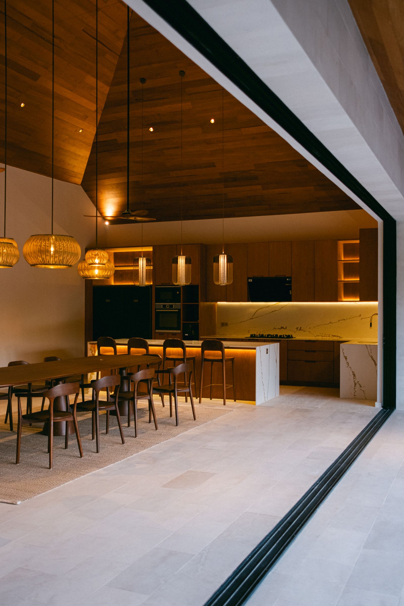 A long timber dining table and open kitchen under rattan pendant lights