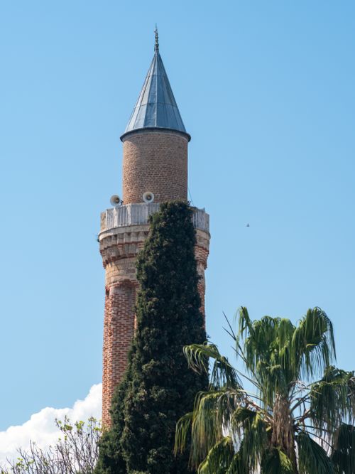 Free A brick minaret tower with a pointed roof and surrounding trees against a bright blue sky. Stock Photo