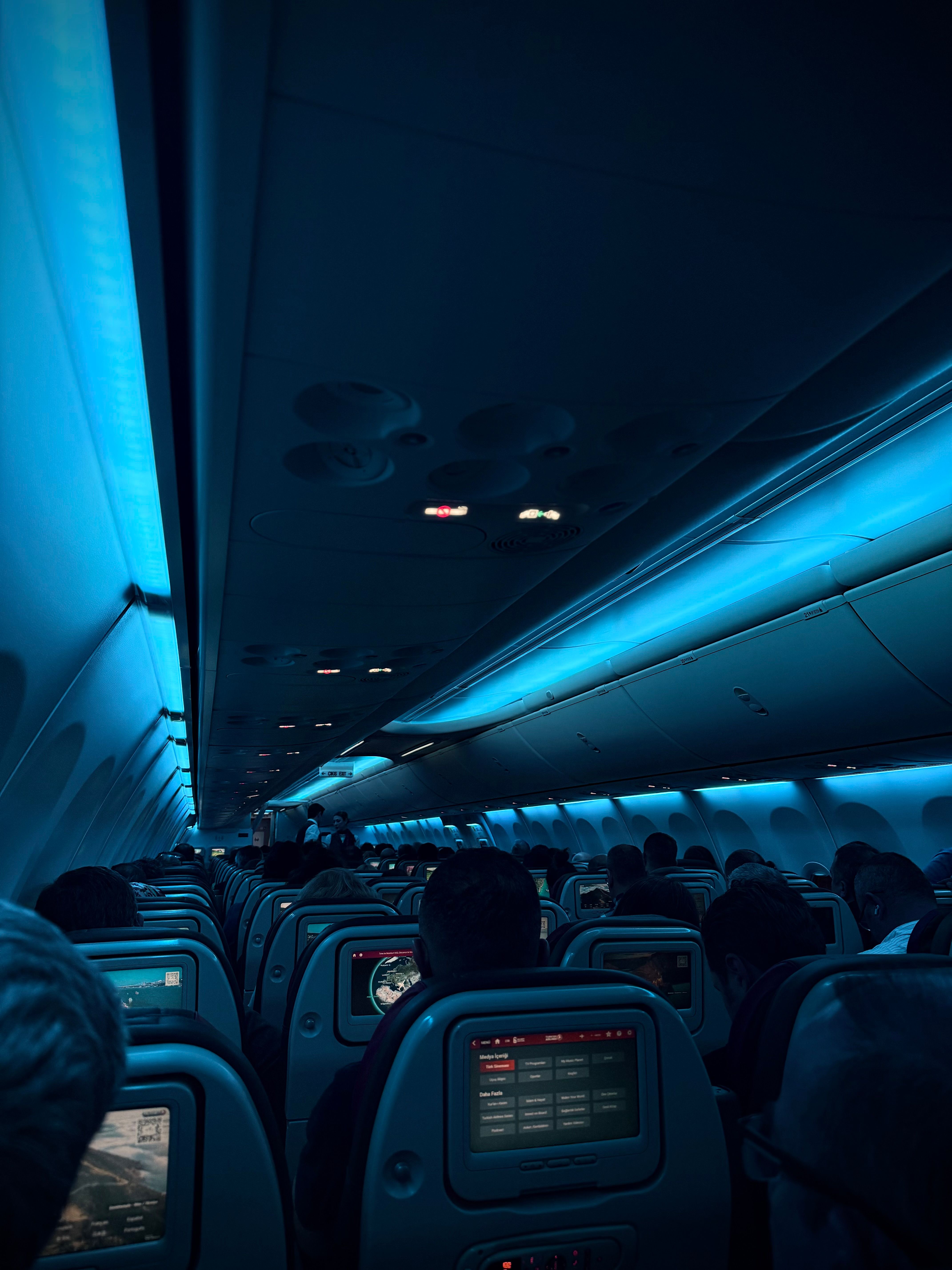 A dimly lit airplane cabin at night with passengers and illuminated screens.