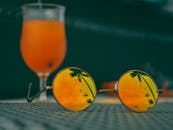 一副充满活力的橙色太阳镜，映出棕榈树的倒影，背景是一杯清爽的鸡尾酒