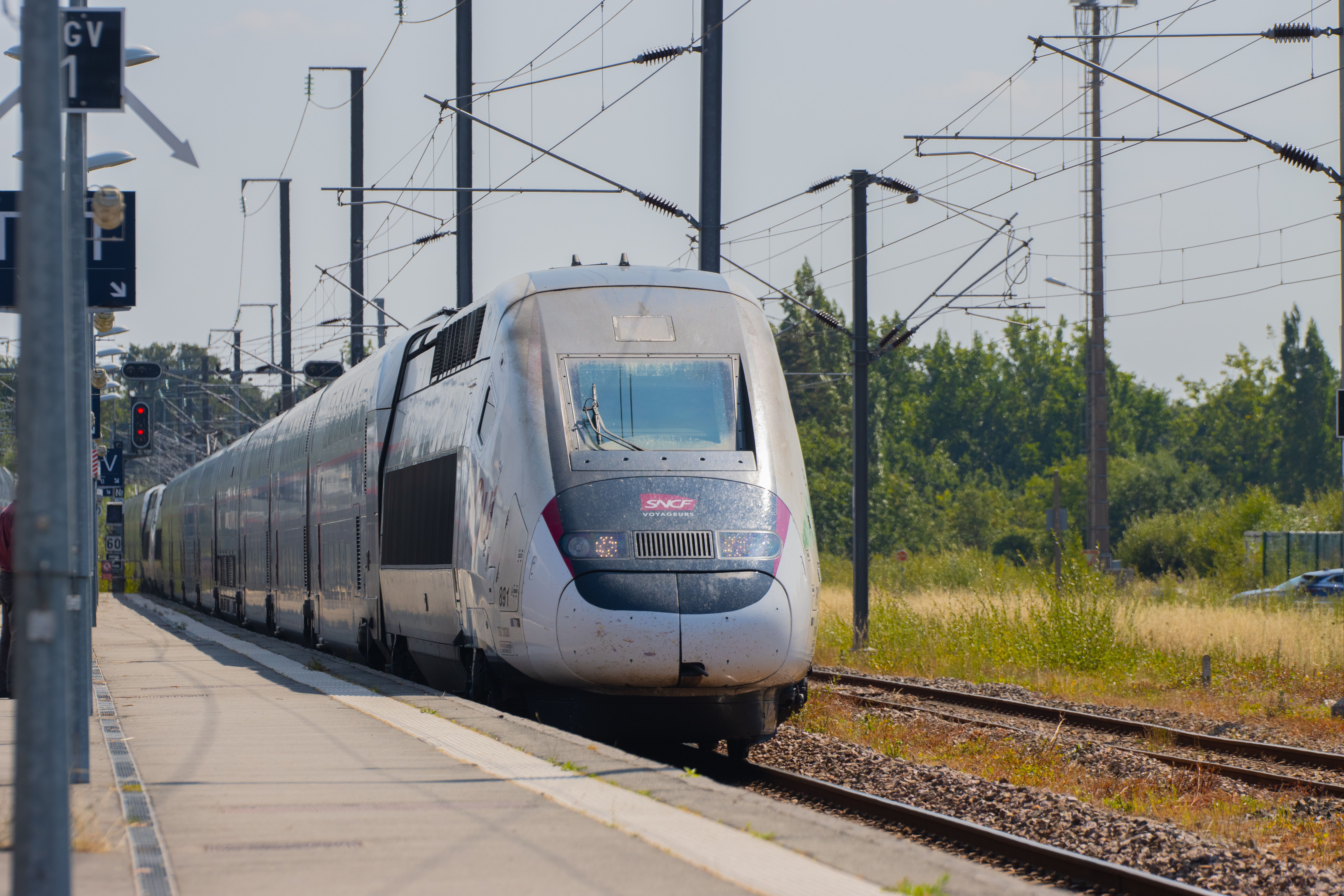 A silver SNCF high-speed train stands beside a railway platform in France, beneath overhead electrical wires