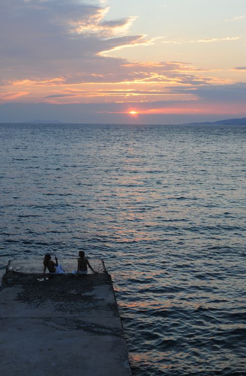 Free Two people sit on a pier watching a serene sunset over the ocean, creating a peaceful ambiance. Stock Photo