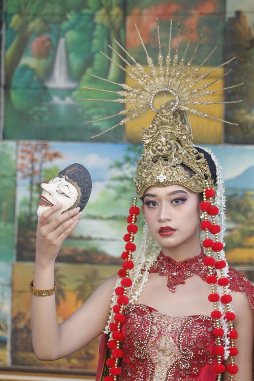 Free A woman in ornate traditional Indonesian attire holding a mask with vibrant background art. Stock Photo