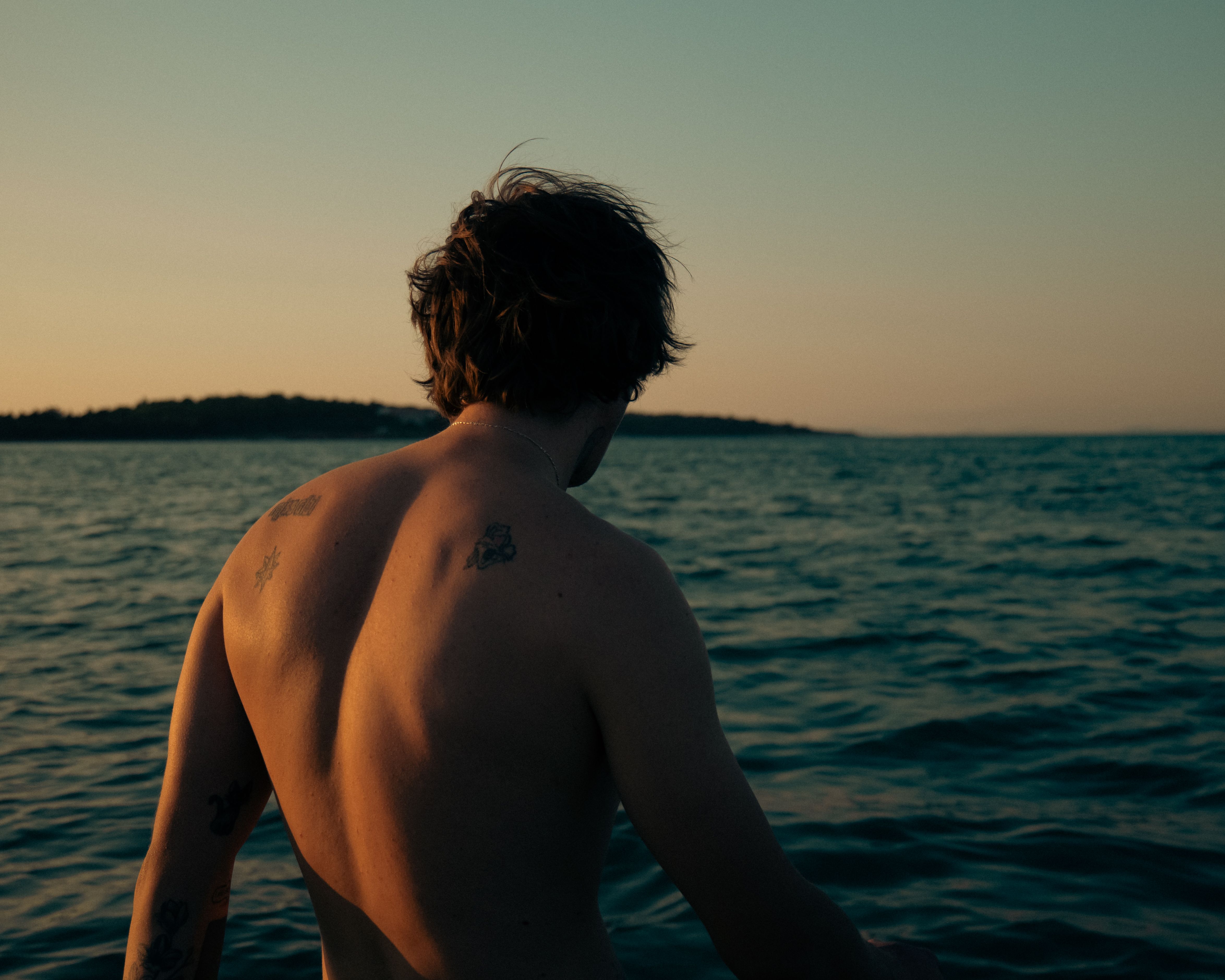 A tattooed man gazes at the calm ocean during a serene sunset, exuding peace.