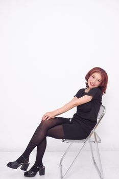 A cheerful woman seated on a chair in an indoor studio setting against a plain white backdrop.