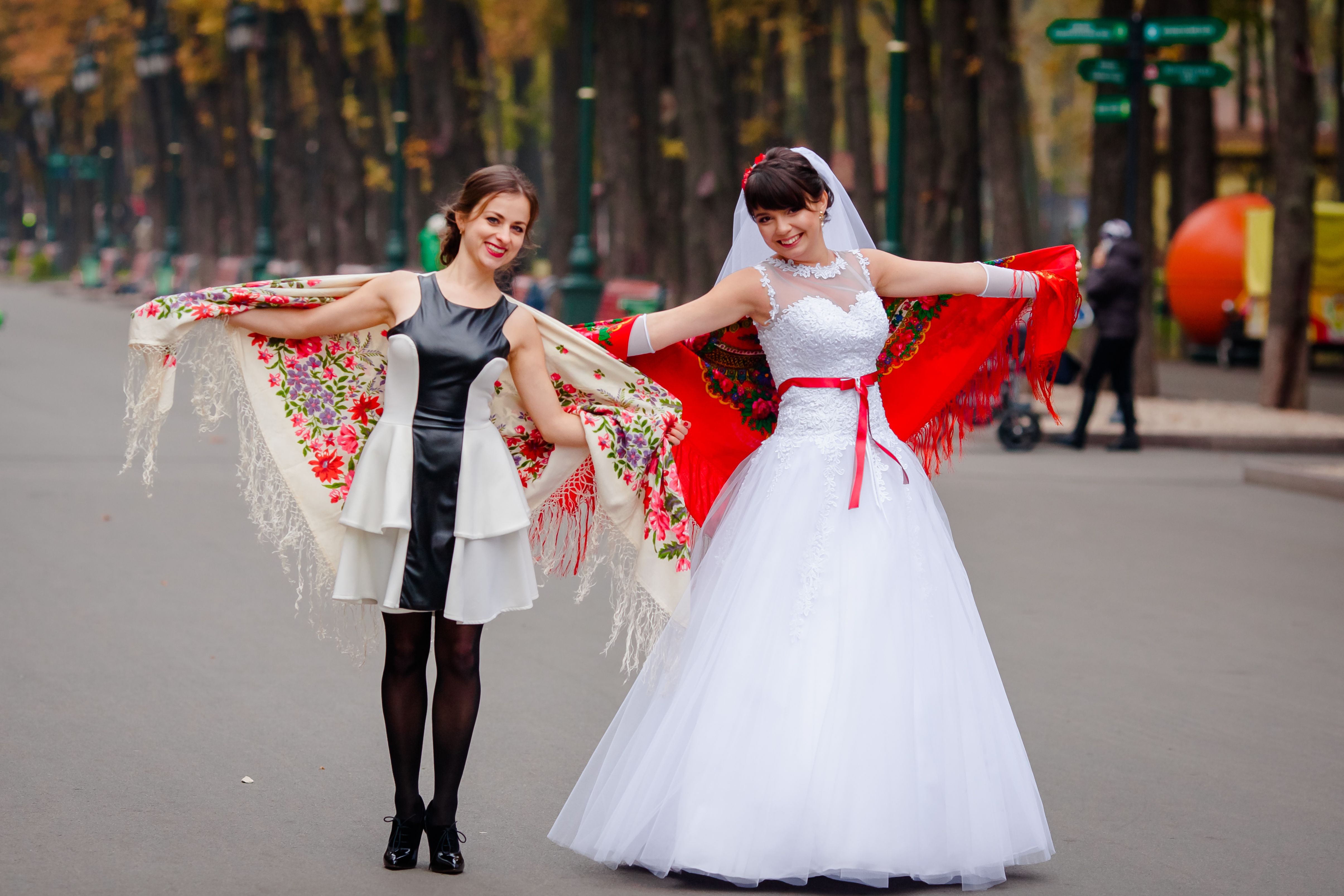 Smiling bride and friend pose in colorful scarves and elegant dresses on a tree-lined street.