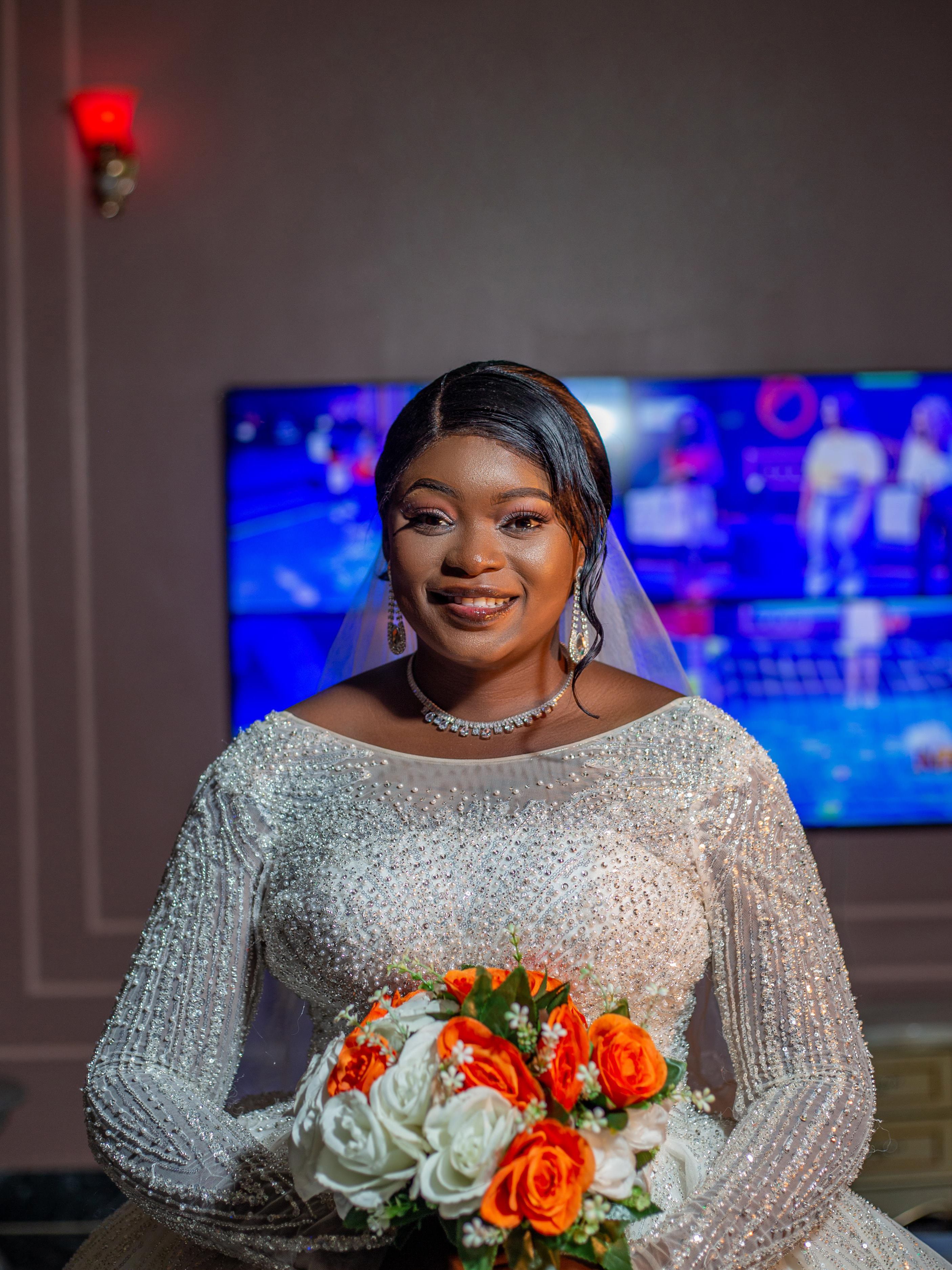 Bride in sparkling wedding dress holding bouquet, smiling indoors.