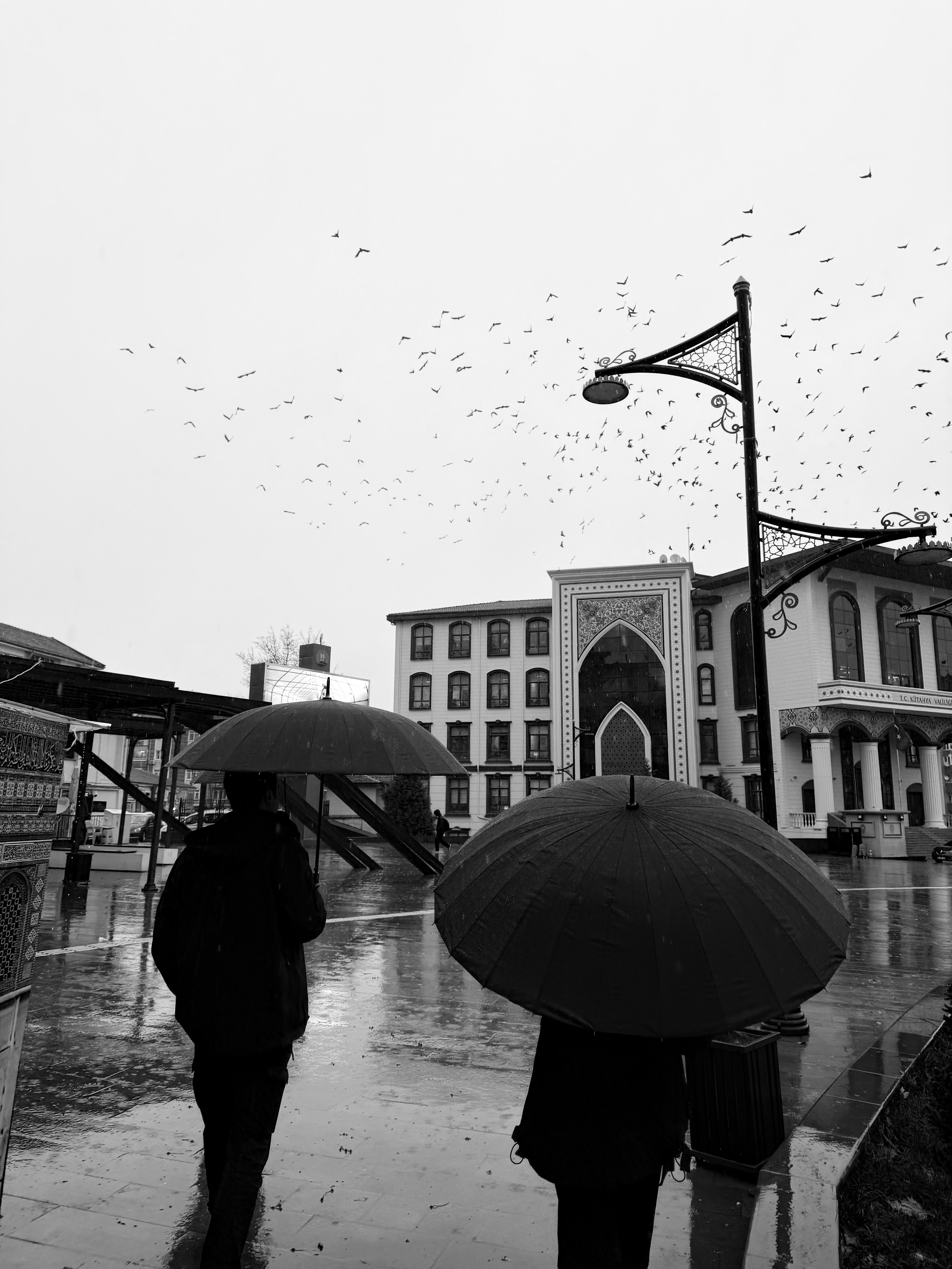 People with umbrellas walk through a rainy city square in Turkey, captured in black and white.