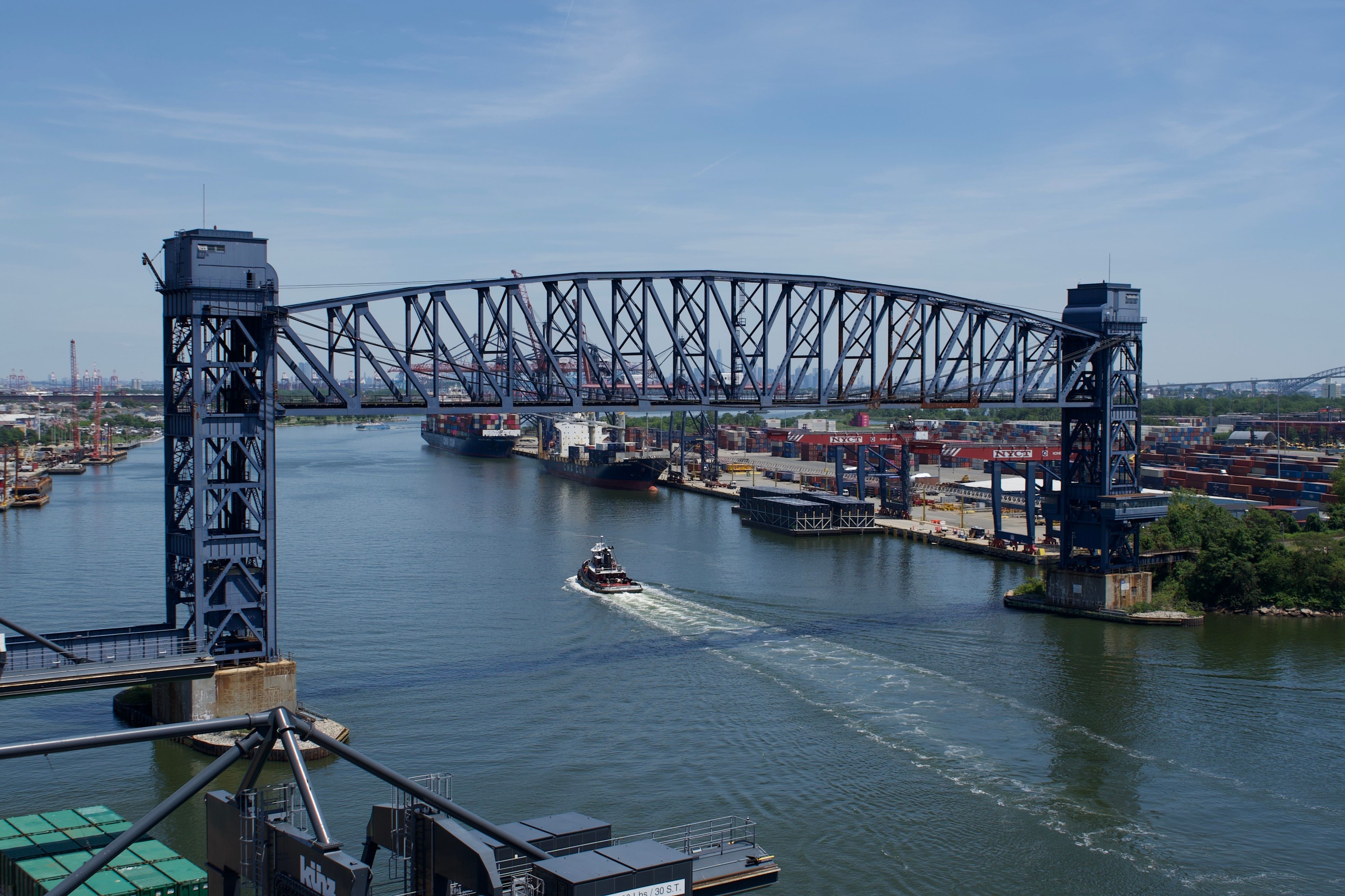Illustrative photograph: A busy industrial harbor with a prominent elevated bridge and a tugboat navigating the waterway.