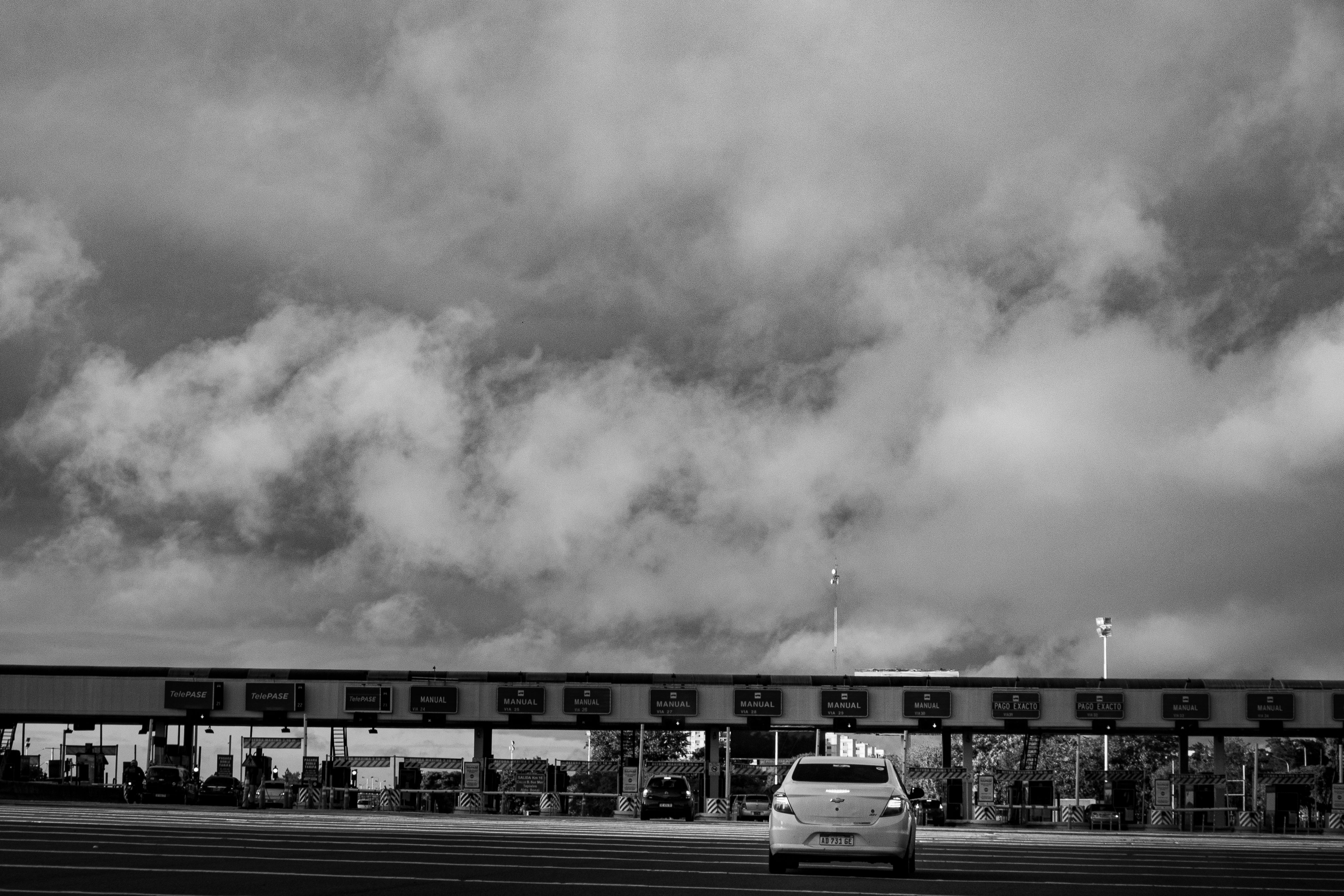 A lone car passing through a toll booth under a cloudy sky in a black and white style.