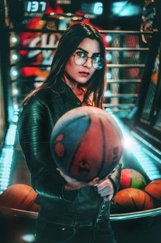 Young woman holding a basketball in an illuminated arcade setting, wearing sunglasses.